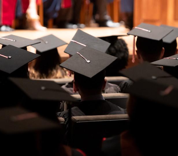 A group of graduates during a commencement ceremony. They are wearing traditional black caps 和 gowns. The setting is Alice Tully Hall with rows of seating filled with the graduates. The angle of the photo suggests it’s taken from the back of the hall, focusing on the back of the graduates' heads 和 their mortarboards, with one in the foreground serving as a focal point.